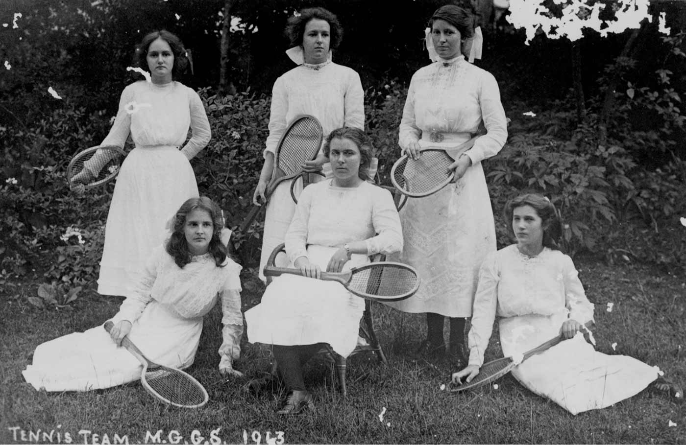 A black and white photo of a group of six women in tennis dresses and holding tennis racquets.