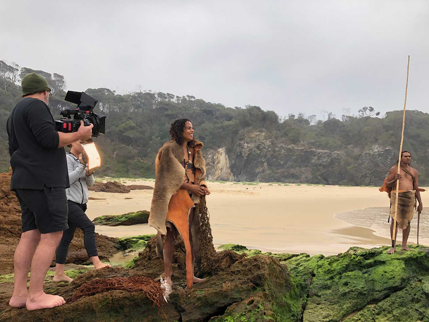 A female actor draped in animal skins stands on a beach with a film crew around her.