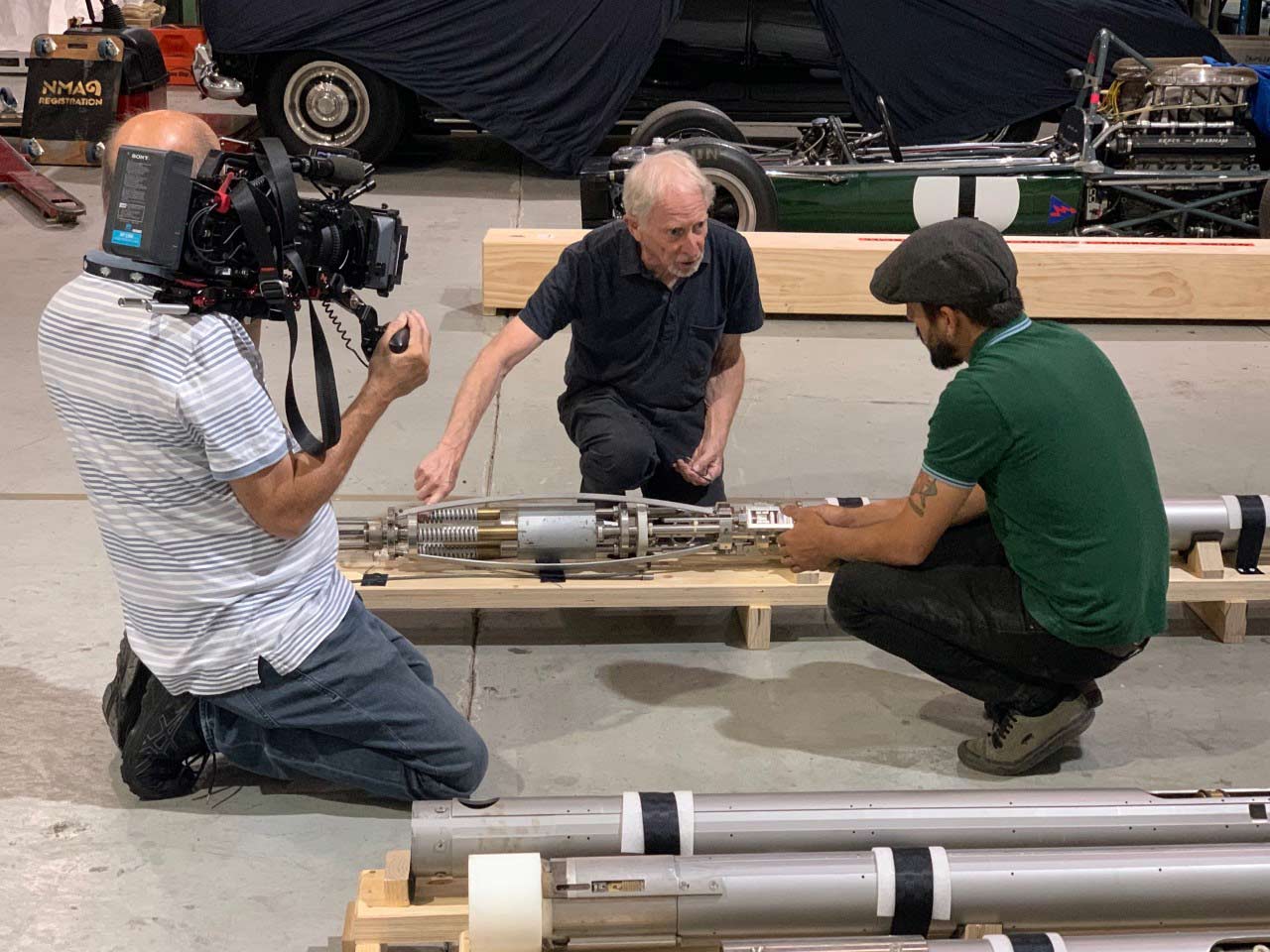A man is filming two other men who are in discussion and kneeling over an ice core drill.