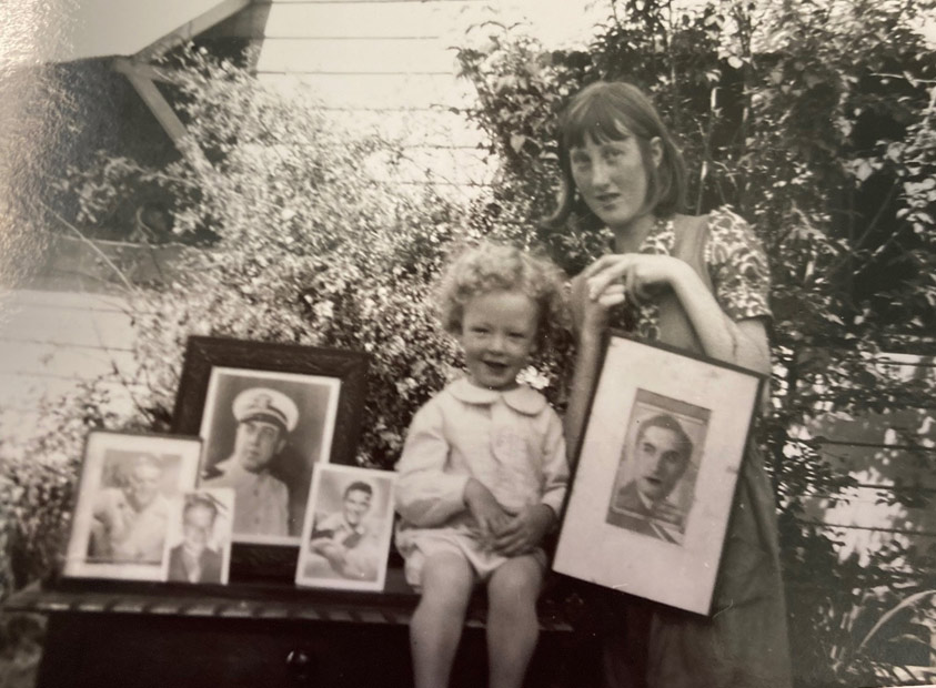 A black and white photograph of a young child seated next to a woman standing, both surrounded by framed photograph portraits of a man.