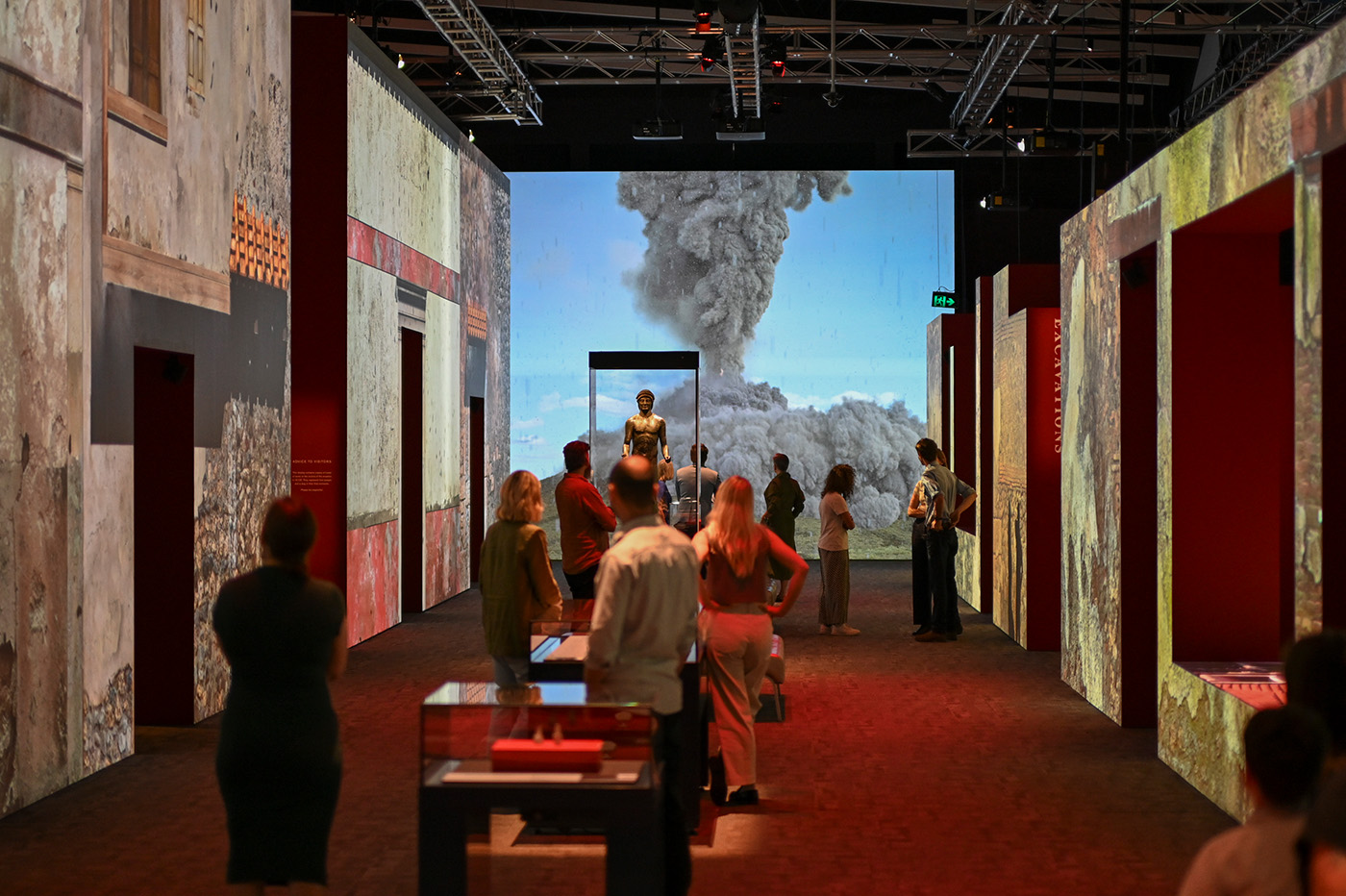 Visitors stand in a large exhibition gallery looking towards a large screen showing a recreation of Mt Vesuvius erupting.