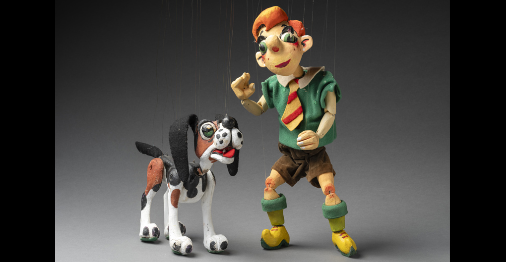 A photo of two string puppets. Right is a puppet of a boy named Nicky, he has bright orange hair and is wearing a green short sleeve shirt, brown shorts and a red and yellow tie. On his legs are green socks and yellow clog-like shoes. Left is a wooden puppet of a dog called Noodle. Noodle has a black, brown and white painted body, felt ears and green felt soles attached to its feet. Noodle has green painted eyes and a red felt tongue. 
