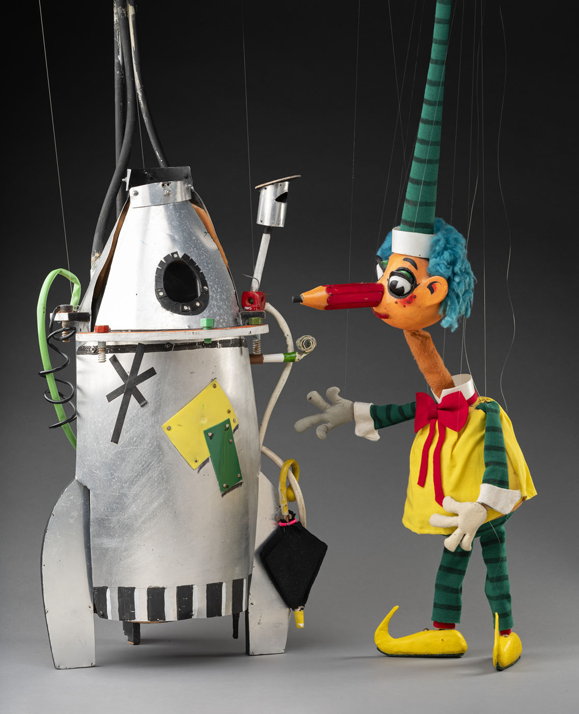 A photograph of Mr Squiggle and Rocket. The rocket is silver decorated with black, orange and white paint. It has green and white tubing and wires, one of the tubes attaches to a felt umbrella. The rocket also has a small circle window with a black frame. Mr Squiggle puppet with blue hair and a pencil for a nose. He is wearing a yellow pinafore with a red bow around the neck, green and black striped sleeves and leggings. He has white gloves, and yellow clog-like shoes that curve at the toes. His nose is a large red pencil with a piece of black oil pastel inserted at one end. Attached to his head is a long tapered green and black striped hat. 