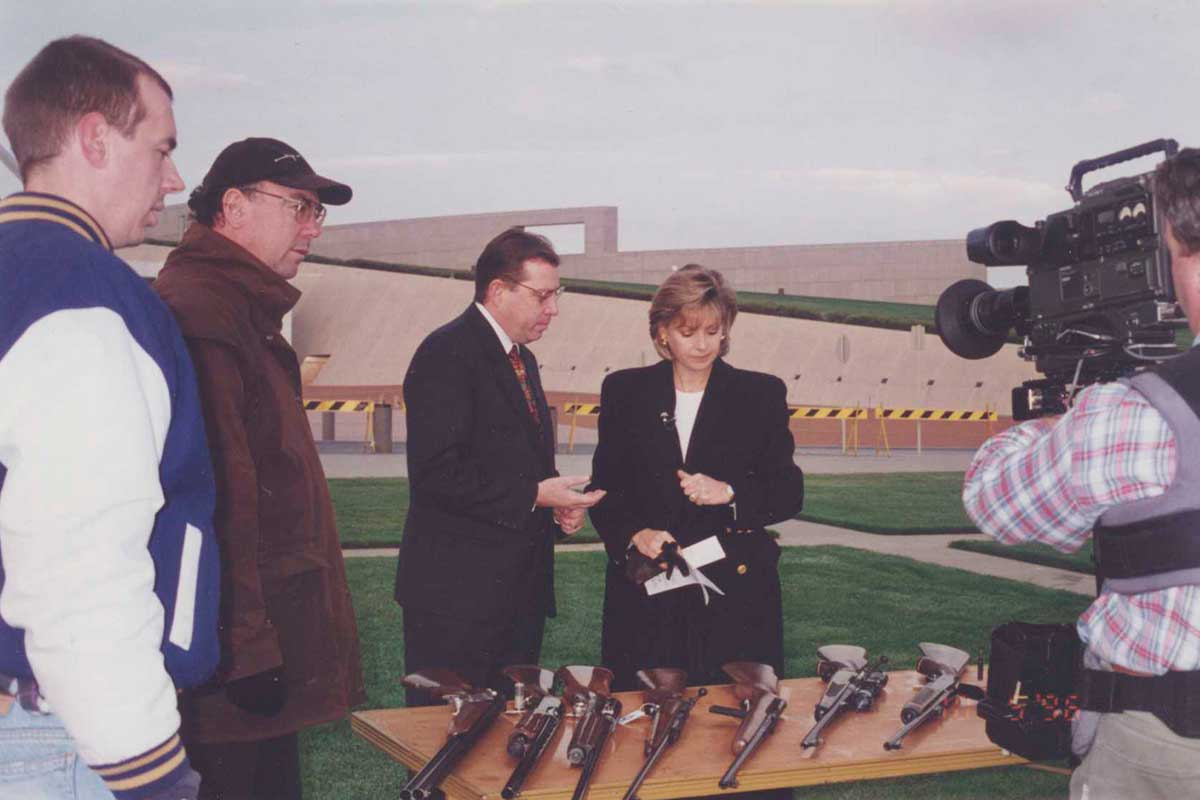A person is filming a group of people looking at several guns on top of a table outside Australian Parliament House.