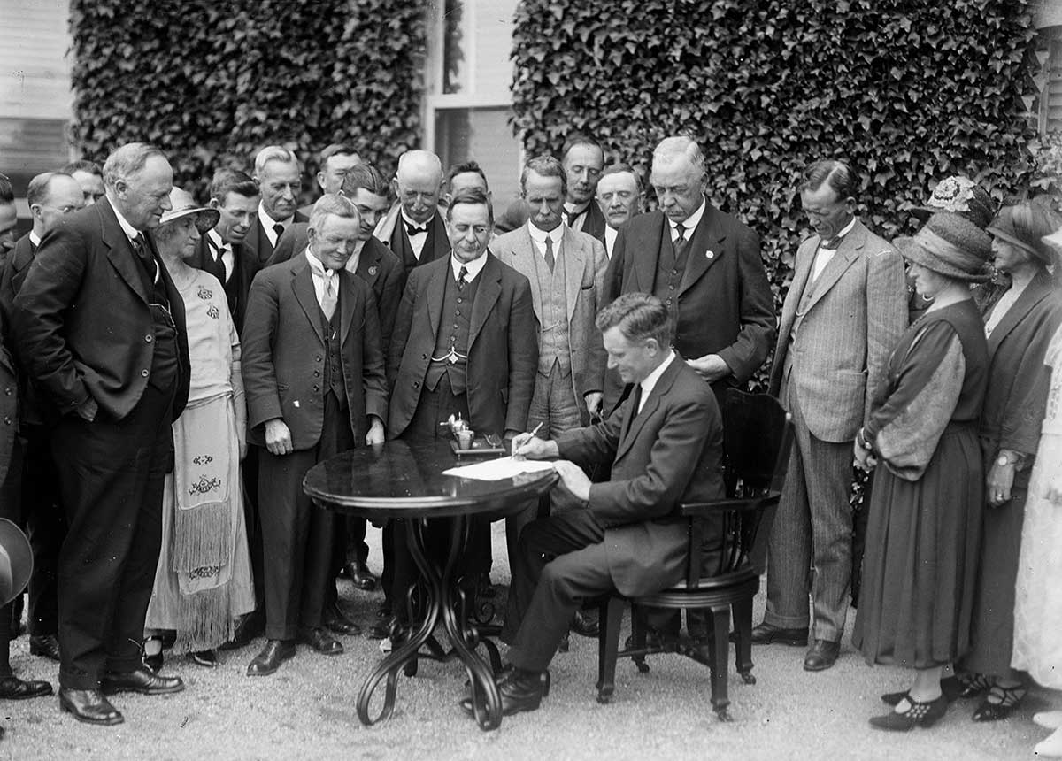 Black and white photo of a group of people watching a seated man at a table signing a document.