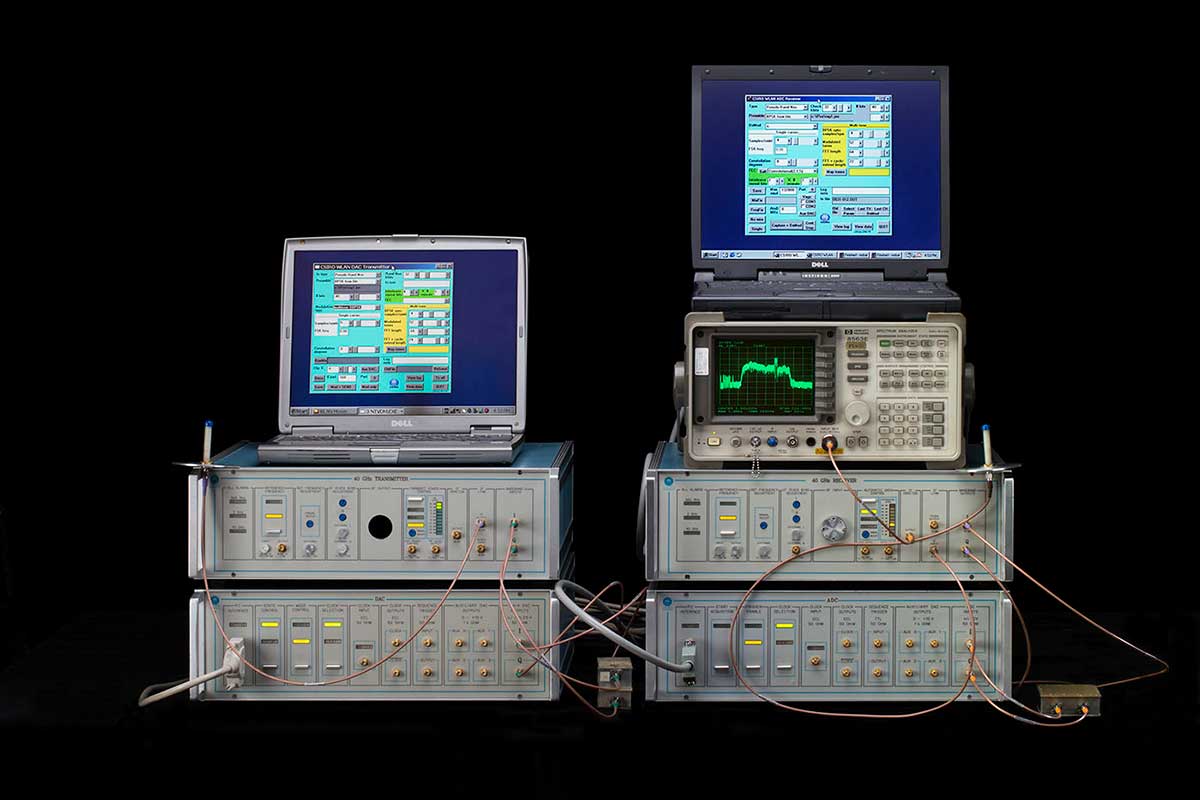 Computer set-up with four grey hardware boxes in two stacks topped by two notebook computers showing data and a spectrum analyser on the right.