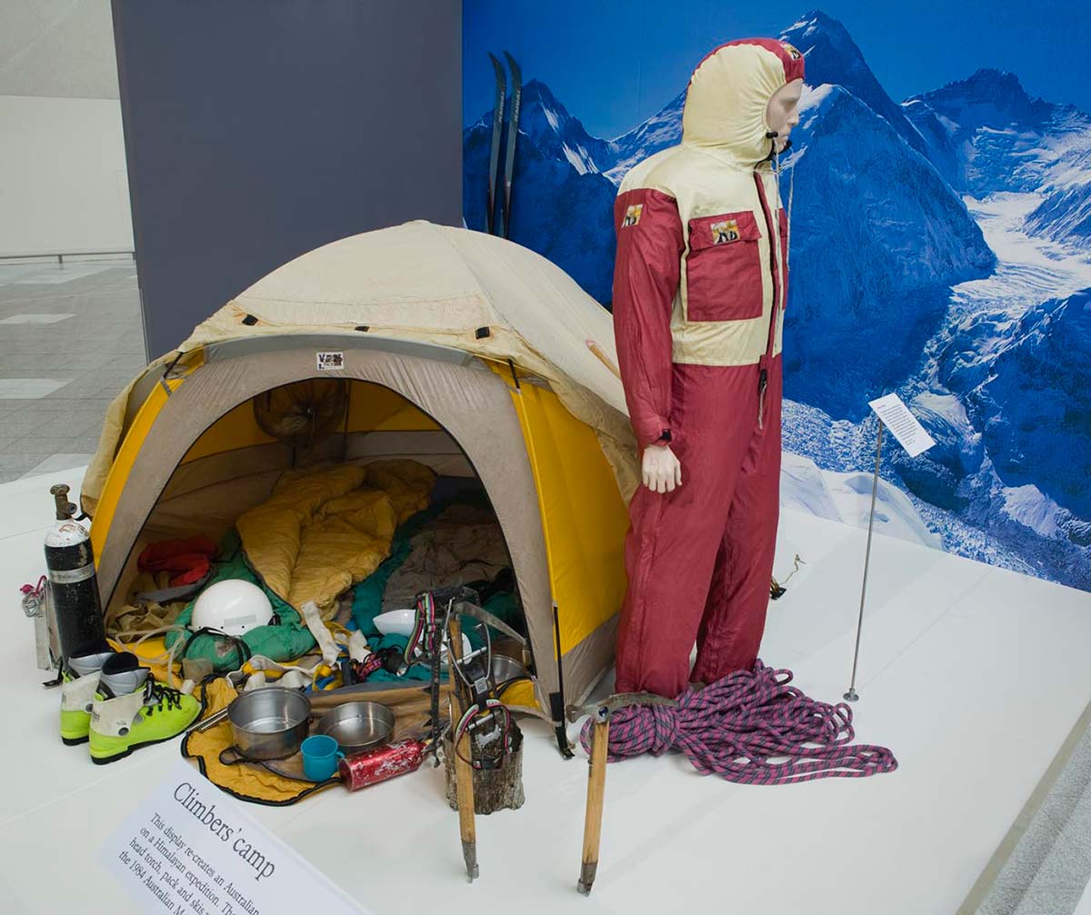 An open tent with camping and mountaineering gear inside and at the front, with a mannequin in a maroon and yellow windsuit at right. An image of snow-covered mountains forms a backdrop.