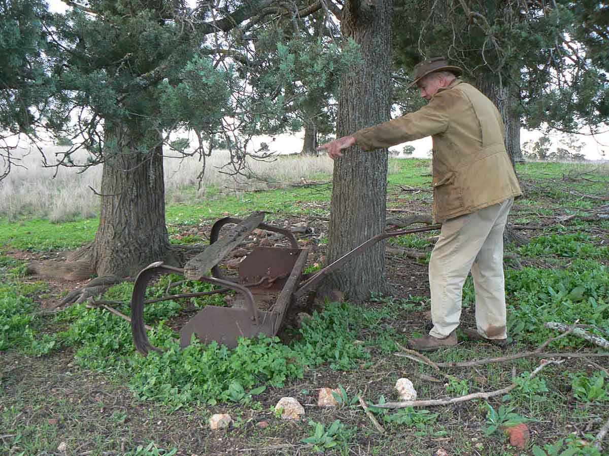 A man standing on farmland at the base of a large tree. He is holding the handle of a piece of rusting farm equipment.