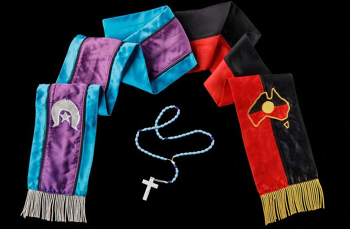 A sash that is half red and black and half aqua and purple. The end of the red and black side has yellow fringe along the edge. There is an Aboriginal flag in the shape of Australia above the fringe. The purple and aqua side has white fringe with a white Torres Straight islander flag.