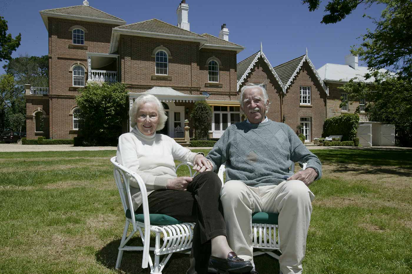 Pamela and Jim Maple-Brown seated on wooden chairs on the grass in front of the main Springfield homestead.
