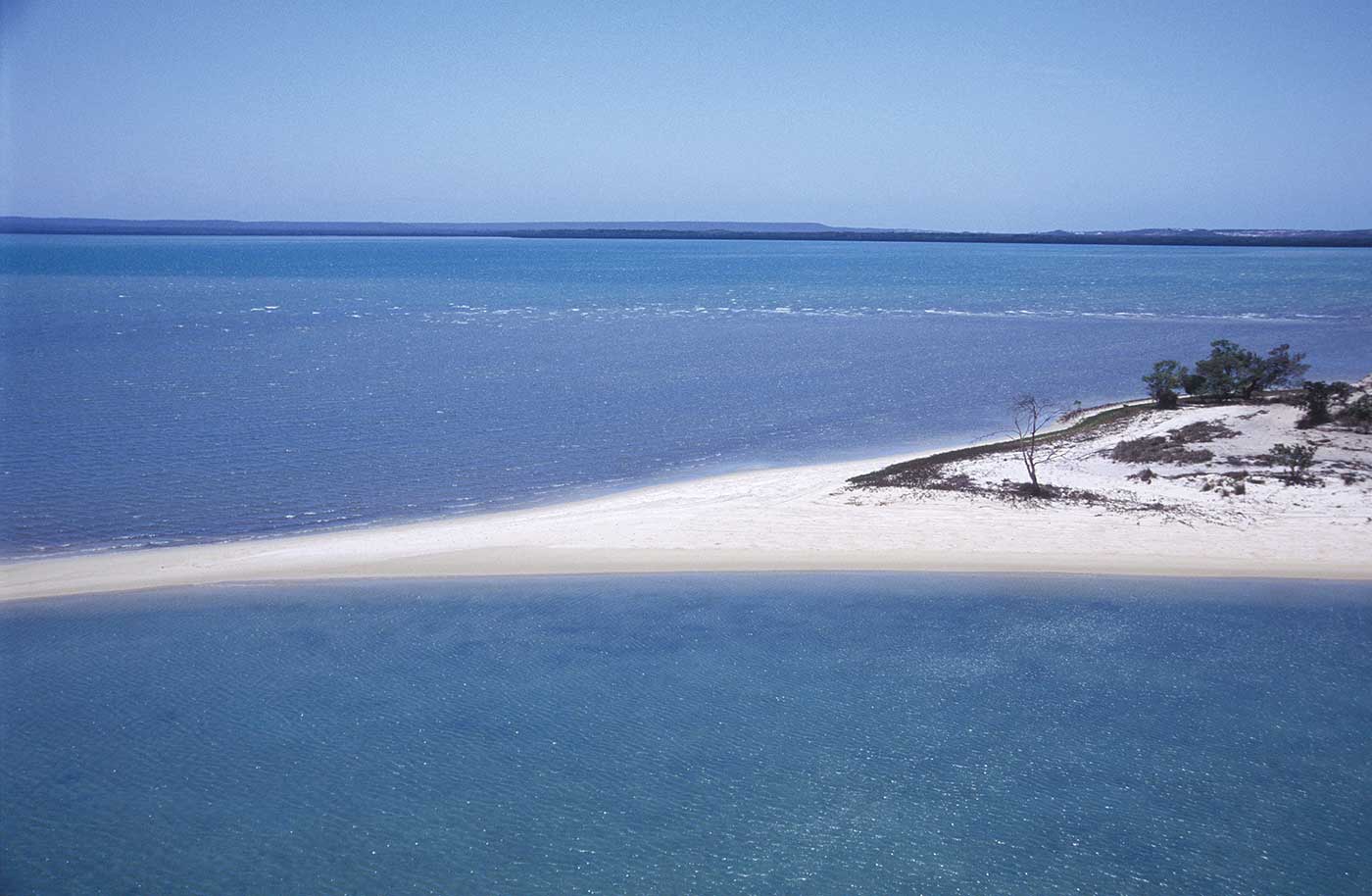 A colour photograph of a sandy spit emerging from a section of coastline. The spit runs more or less across the middle of the photograph. It begins at the right and narrows to the left. There is ocean on either side of it. Some sparse vegetation grows on the base of the spit. On the horizon appears to be some more coastline. The ocean is a rich blue, while the sky overhead is a pale blue.