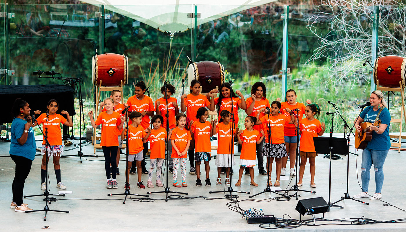 A group of children and adults, wearing orange t-shirts marked with the words 'Four Winds', perform on stage.