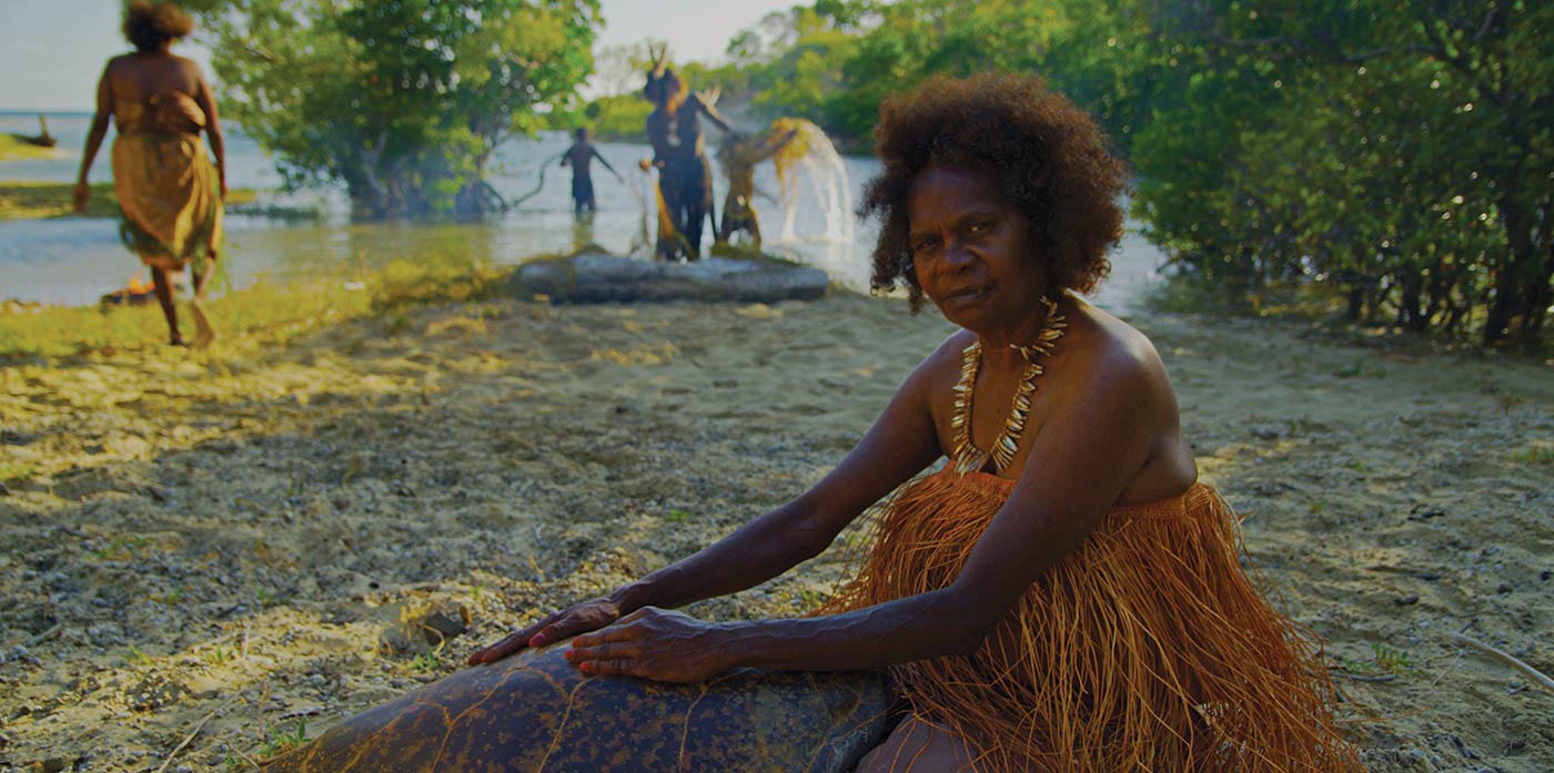 A woman sits on stony sand with her hands placed on a large turtle shell. She is wearing a grass top. There are people in the background working with nets in the shallows.