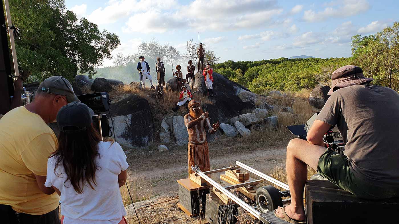 Film set featuring a scene with a ship's captain and soldiers posing on rocks with Indigenous people. The camera focuses on an Indigenous man who stands and gestures with his hands.