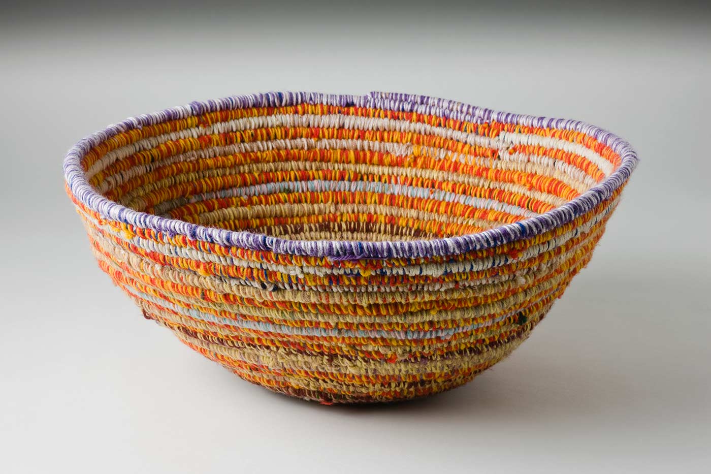 A circular coiled multicoloured yarn and plant fibre basket. The plant fibre in the very centre of the basket is covered with orange-green yarn, followed by horizontal alternate yarn coverings over the fibre. The alternate yarn colours are brown, brown-blue, yellow-blue, yellow-brown, tan-cream, tan-brown, cream-pink, red-yellow, yellow-tan, light blue-light pink with various combinations of red, yellow, tan and white up to the top coil. The top plant fibre coil is covered by white-purple yarn.