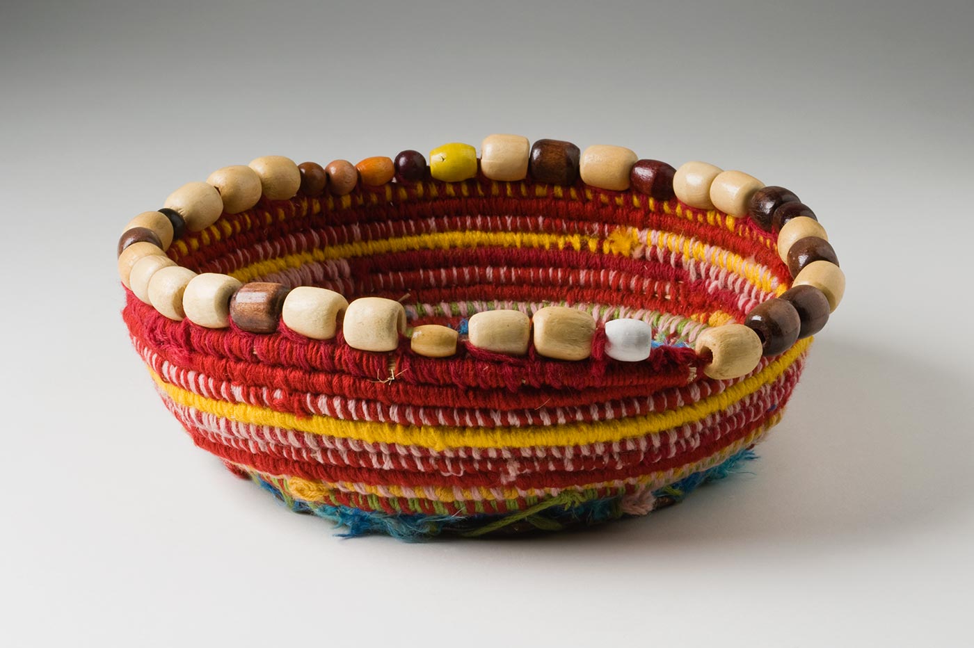 A shallow circular coiled red toned yarn and plant fibre basket with a metal base and bead trim. The base is made of a