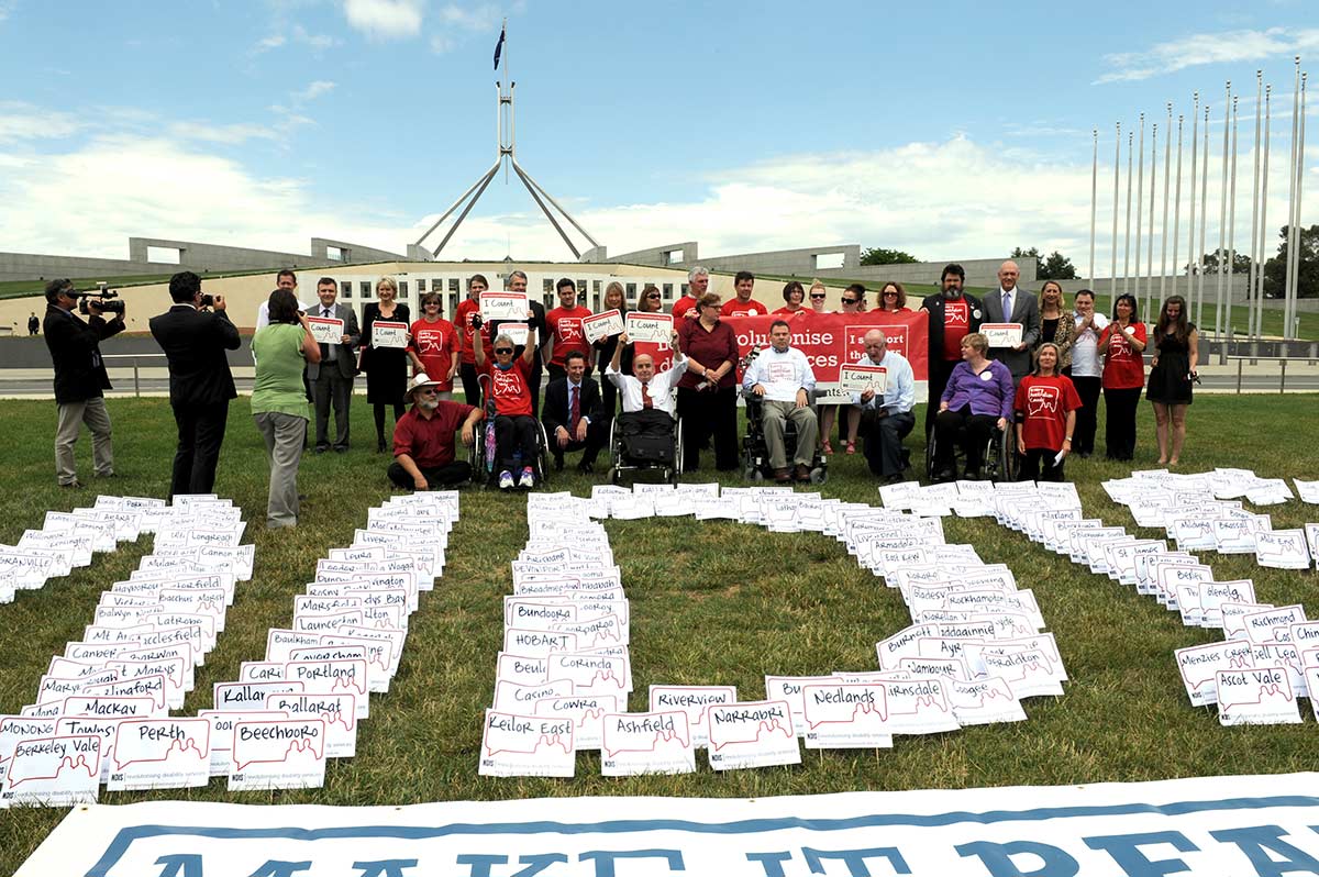 A group of campaigners and the media are gathered on the lawns of Parliament House in Canberra. The campaigners, some in wheel chairs, are holding up placards. Signs with cities, towns and districts across Australia in the formation of NDIS lie on the grass in front of them..