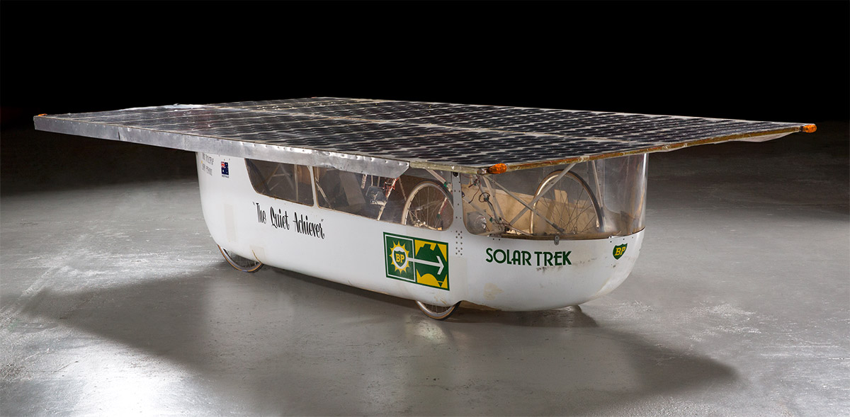 A solar-powered vehicle clad in a white fibreglass, featuring a large flat rectangular array of solar panels mounted on the roof, bicycle fittings including wheels and frame to the interior, and the text