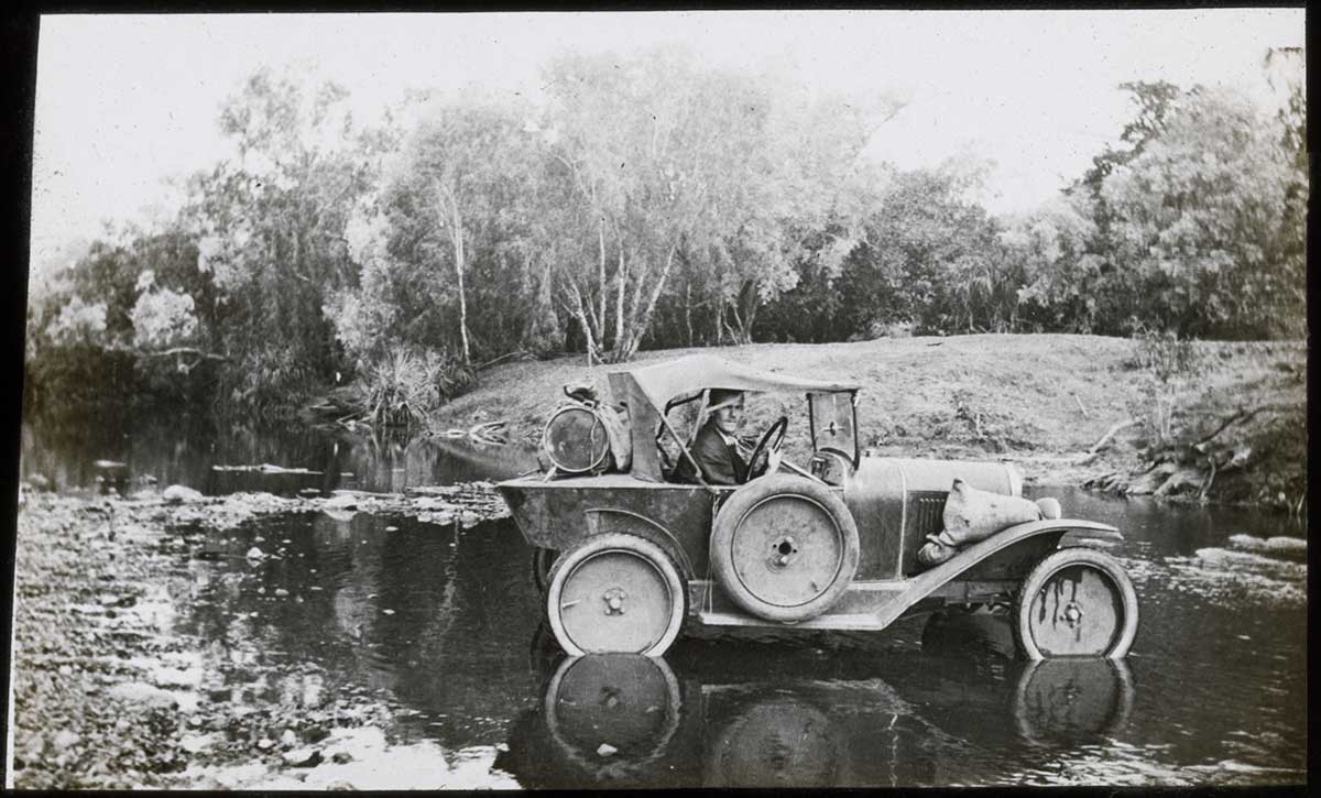A black and white photograph of a man driving an automobile across a shallow river or creek in bushland. There is cargo on the back of the automobile, a spare tyre attached to the driver's side, and an unidentified sack on the front right fender.