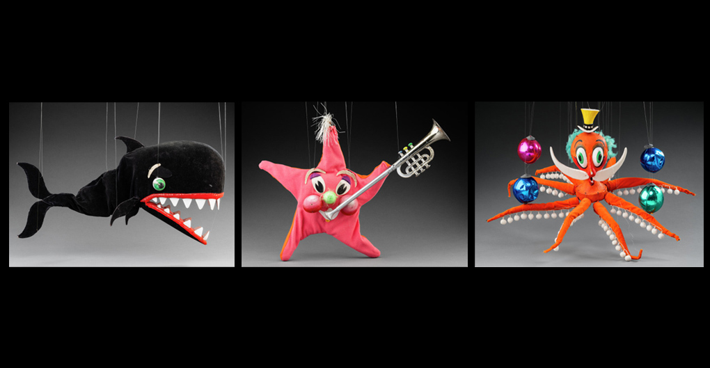 Left: A marionette of a black whale with green eyes. The whale’s mouth has red lips and white triangle shaped teeth.  Middle: A marionette of a pink and orange starfish with a green nose and two large red cheeks. At the top of the starfish is a tuft of white ribbons that look like hair, and in its left hand is a silver and gold trumpet. Right: A marionette of an orange octopus and a white moustache wearing a yellow hat. Accompanying the puppet are plastic balls in blue, green and pink. 