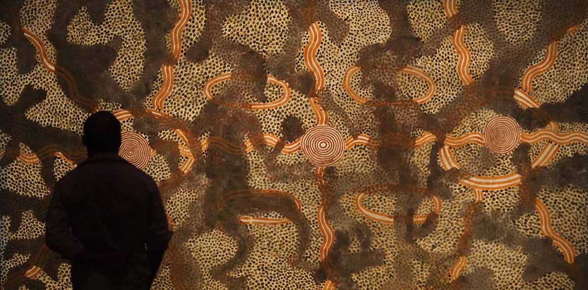 Silhouetted man standing in front of painting featuring three concentric circles, centre bisected by shadowy, wavy crossed lines and figures. Others surrounded by oval shape, four wavy lines in diagonal crosses from ovals. Four other ovals around centre circles. White ground with patches of black and yellow dots, and red dots on black.