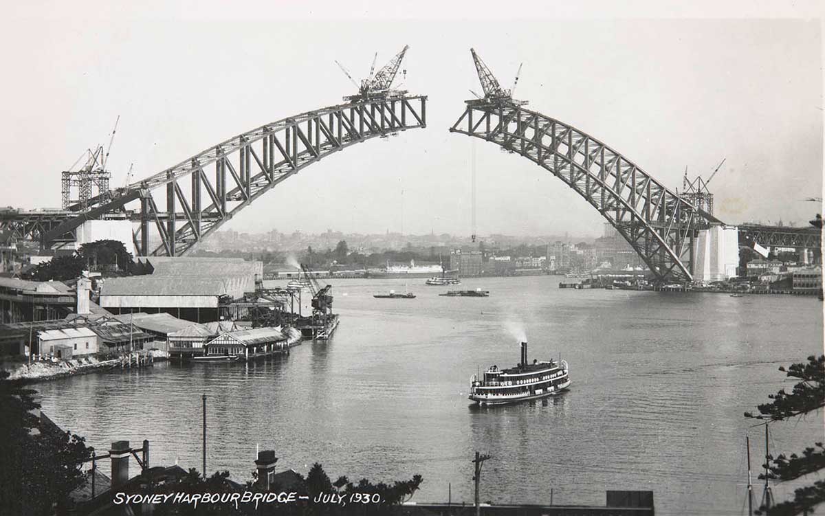 Photo taken from the north shore showing the two sides of the arch under construction. They have yet to meet. Large cranes are perched on top of each end of the spans. A ferry can be seen in the foreground.