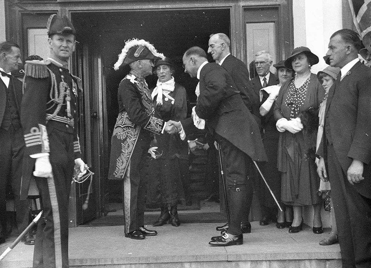 Man of middle height wearing elaborate uniform and plumed hat and shaking hands with man in tails. Other similarly dressed people stand on either side, including senior naval officer in full dress uniform.