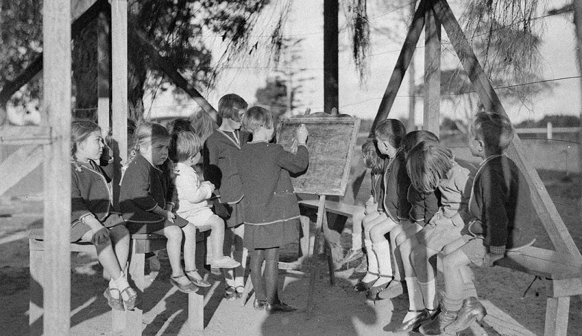 Black and white photo of a group of children around a blackboard.