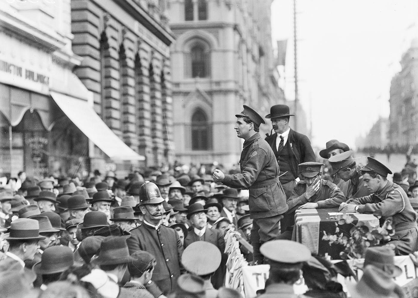 Black and white photo of soldiers campaigning in front of a crowd of men and a police officer.