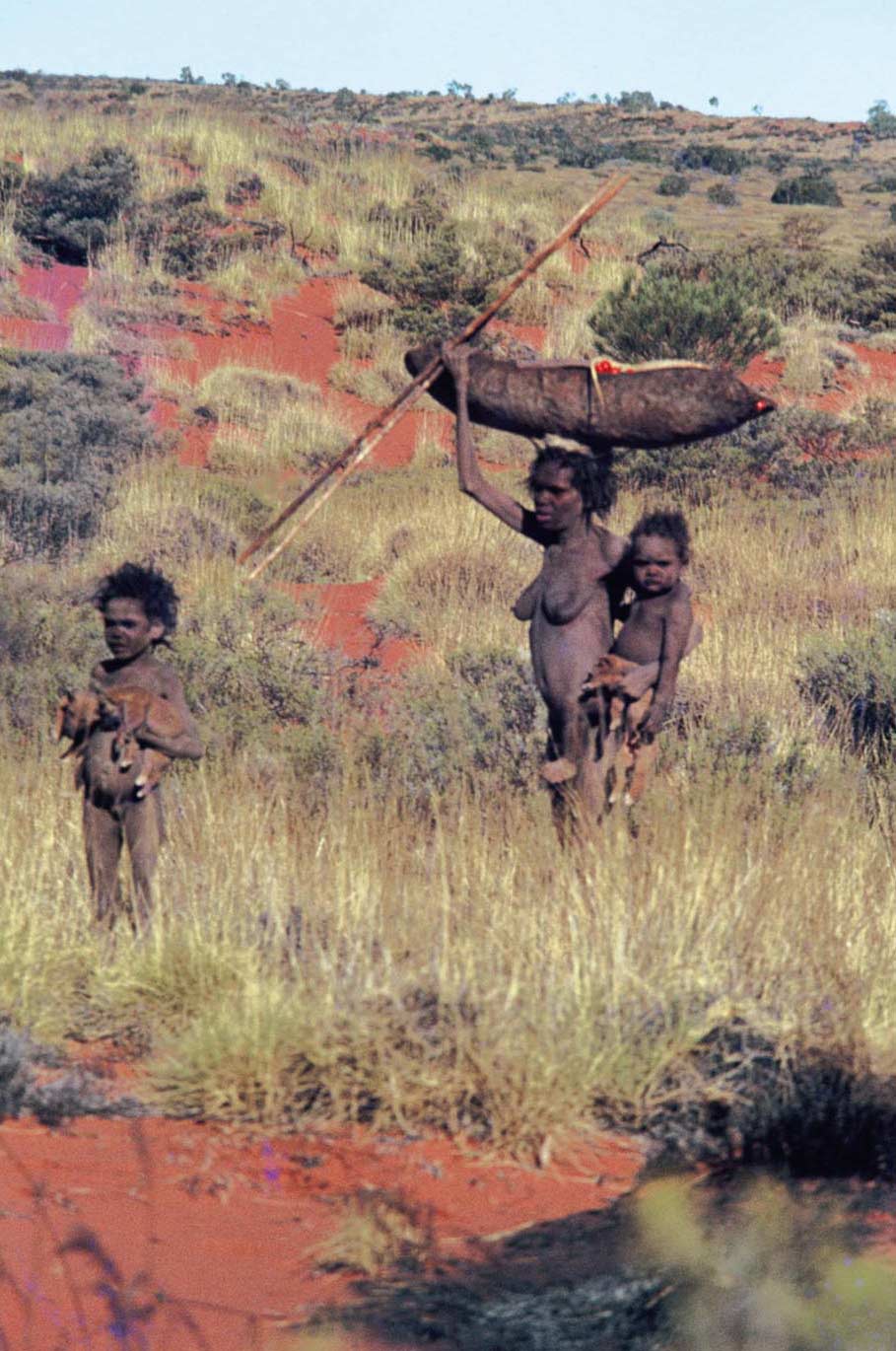 A woman standing in the bush with a wooden vessel on her head. She is carrying a baby and a young child is standing nearby and holding what appears to be two dingo puppies.