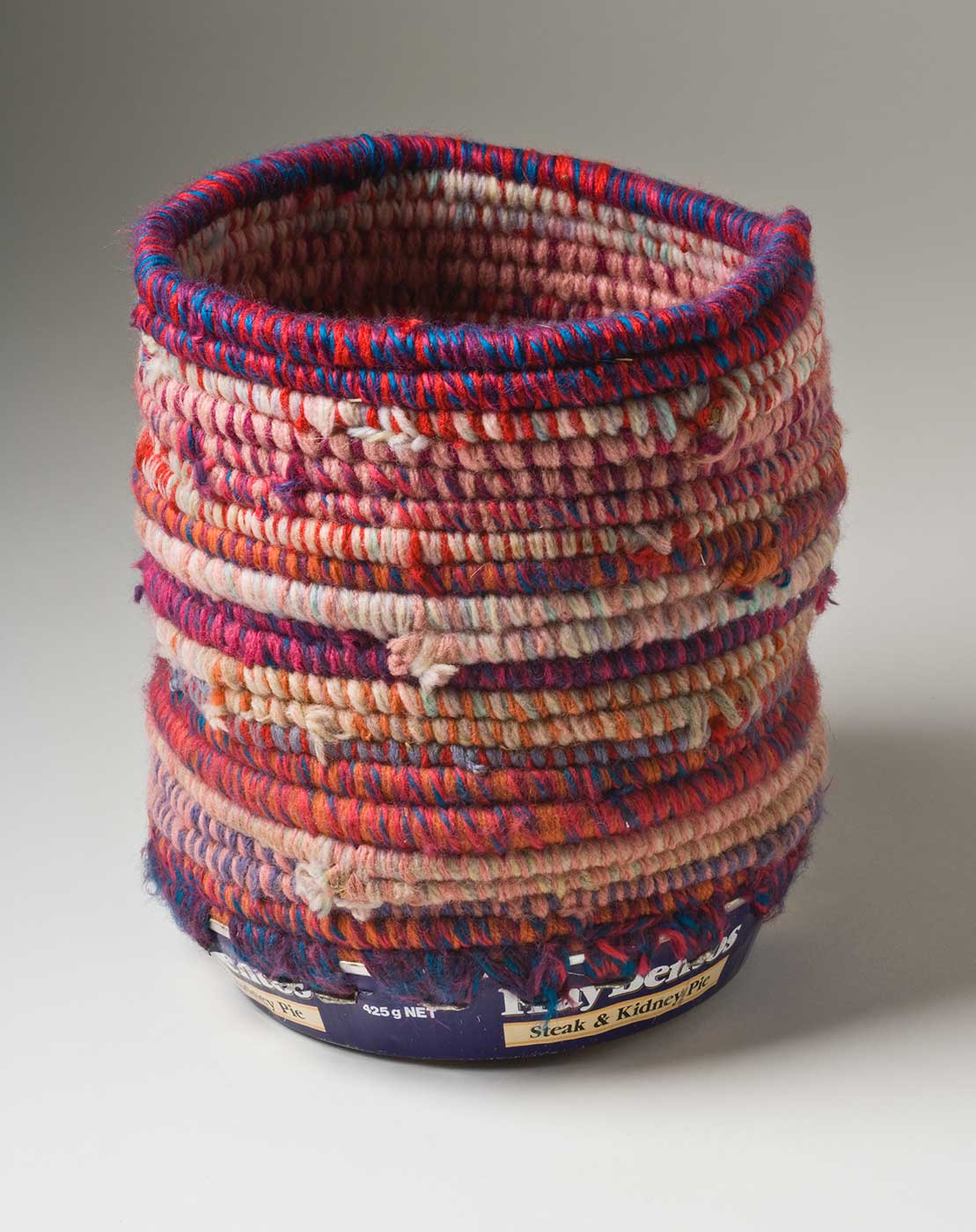 A cylindrical coiled multicoloured yarn and plant fibre basket with a metal base. The coiled yarn is in colours of blue, orange, red and purple with a dominant colour of pink. The base of the basket is a blue and white coloured metal tin with burn marks to the bottom which includes the text 'Fray Bentos / Steak and Kidney Pie'. Yarn is used to attach the base to the top section through holes that have been punched in the tin.