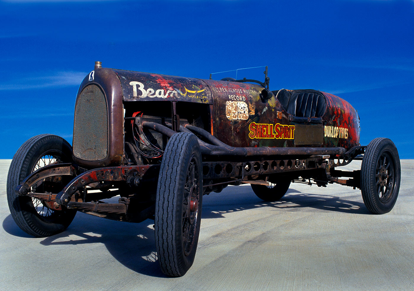 A four-wheeled open seater car with a long engine cowling, boat tail and low glass windscreen. 'Bean' has been painted in white on the top right of the bonnet cover. The words 'Shell Spirit' and 'Dunlop Tyres' are visible on the right side of the car's body.