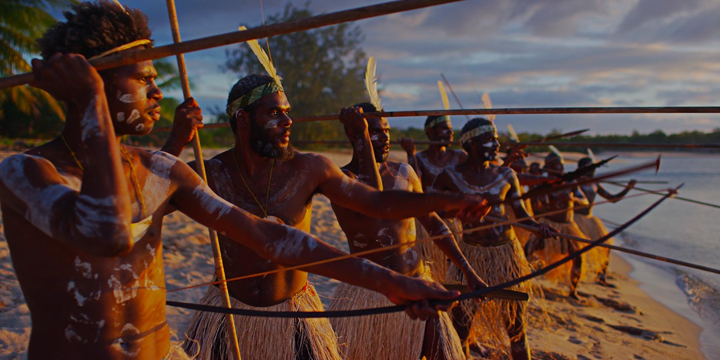 A group of warriors holding spears and ready to attack as they look out from a beach. They wear grass skirts and white feather headdresses.