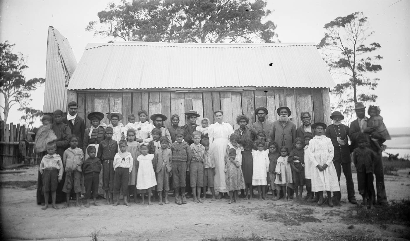 Black and white photograph showing a group of Aboriginal men, women and children standing in front of a slab hut.