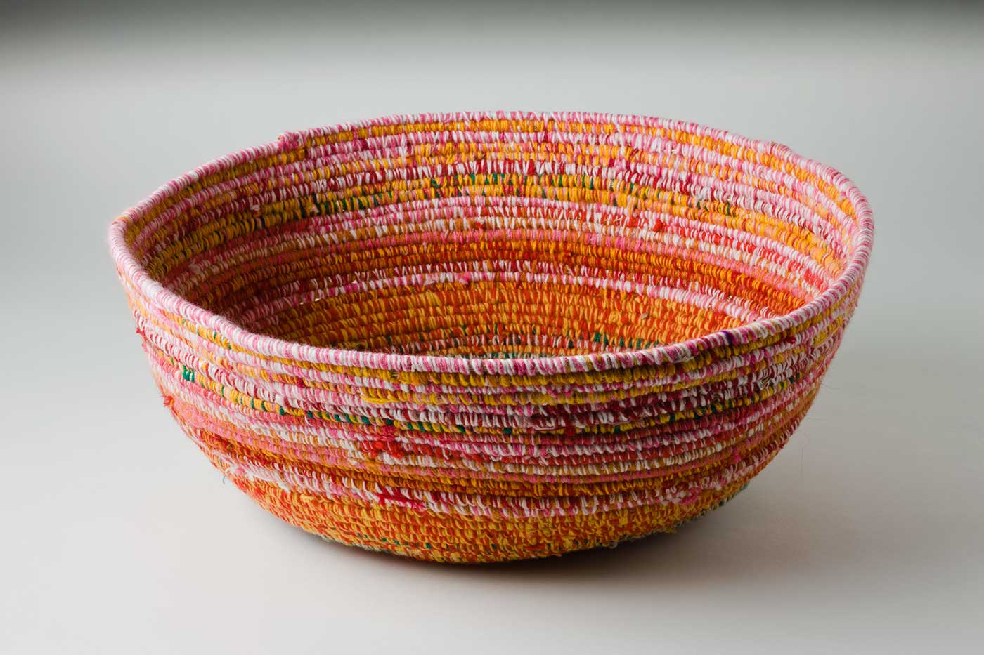 An oval coiled multicoloured yarn and plant fibre basket. The plant fibre in the centre of the basket is covered by brown-blue yarn, followed by horizontal alternate yarn coverings over the fibre. The yarn colours are green-yellow, red-yellow, red-white, red-green, green-white, pink-white, red-brown, pink-brown, and yellow pink. The top plant fibre is covered by pink-white yarn.