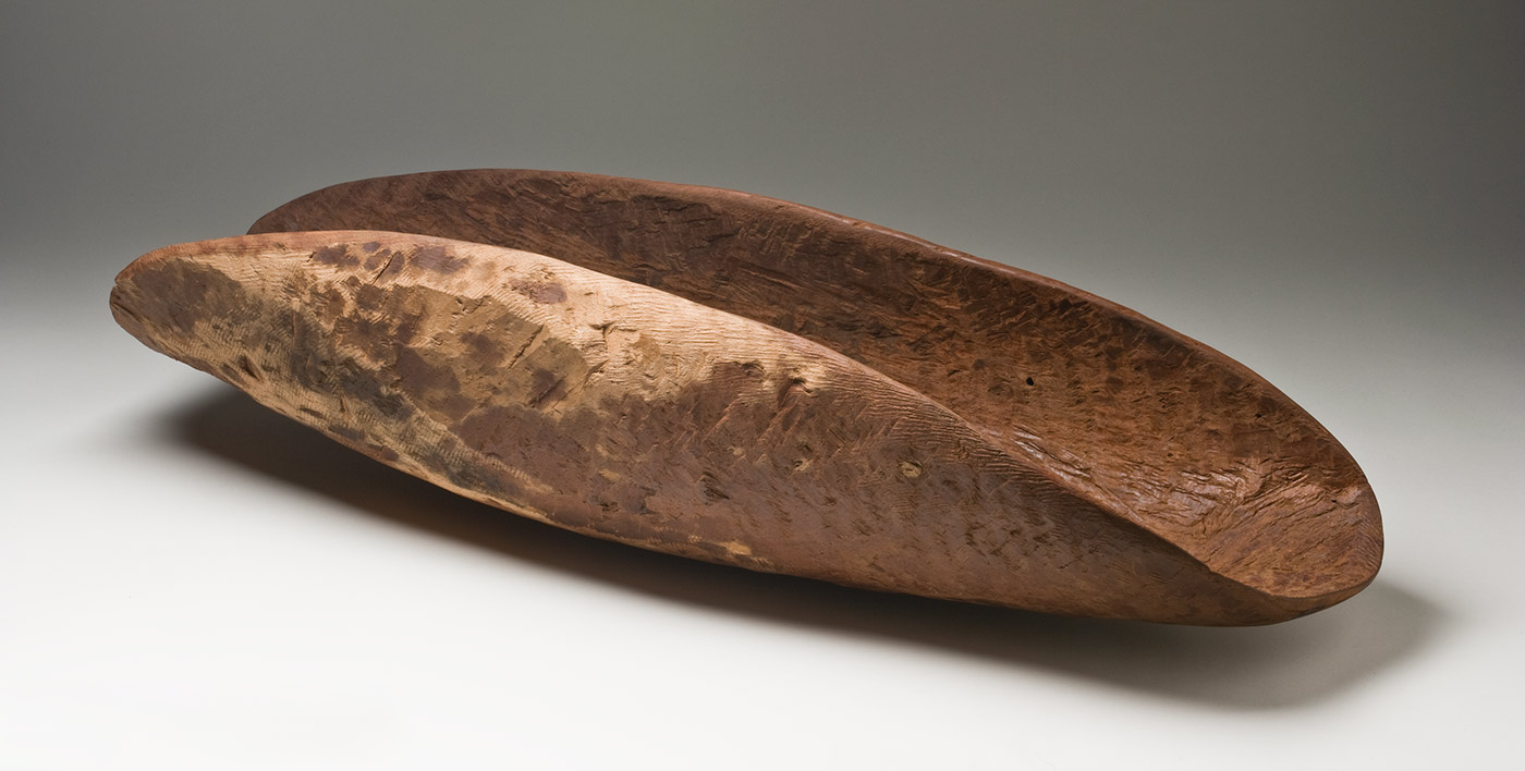 A red-brown wooden concave oval container with dark patches on the sides of the outer surface. The inner surface is rough with the tool marks and there are short parallel grooves along one end of the inner surface. The outer sides are a light beige colour.