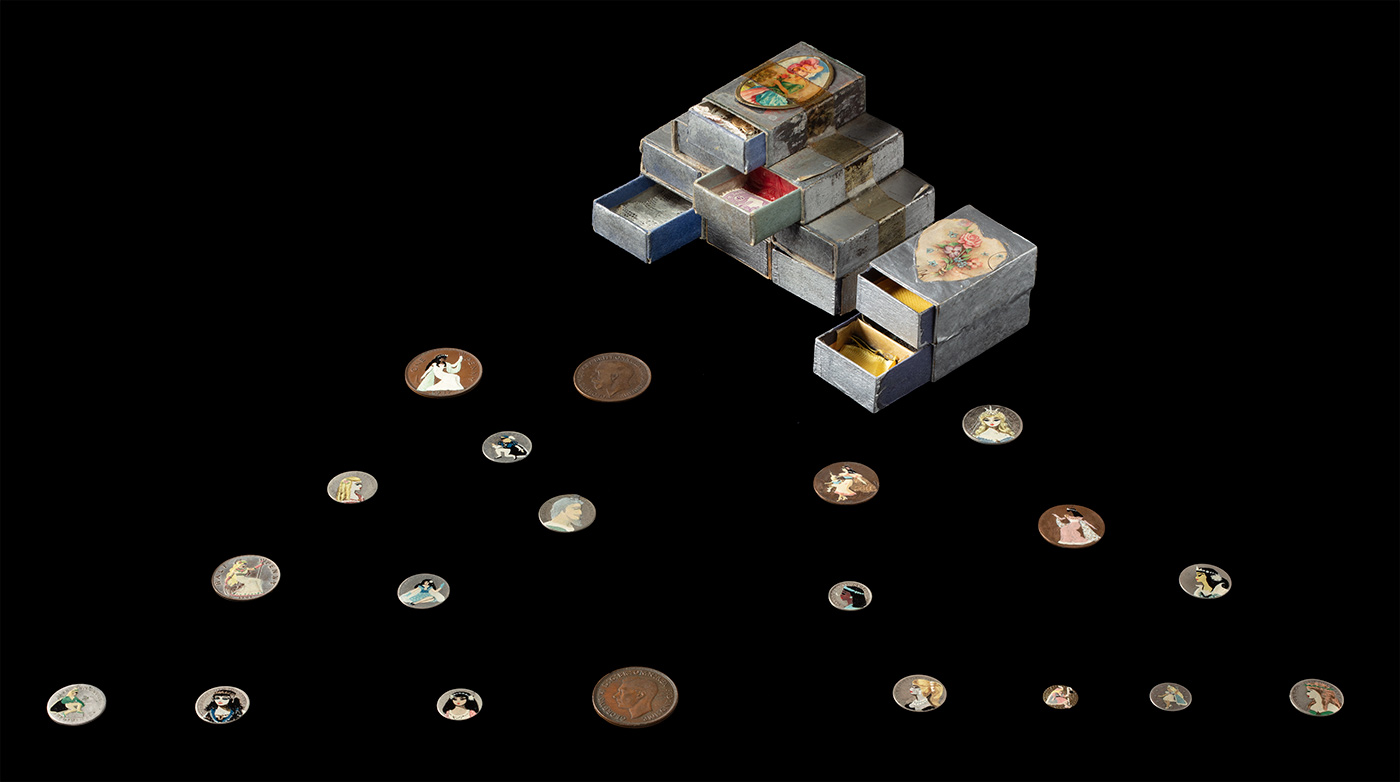 Collection of antique coins with hand-painted images on their surfaces, and adjacent to a stack of decorated matchboxes on display.