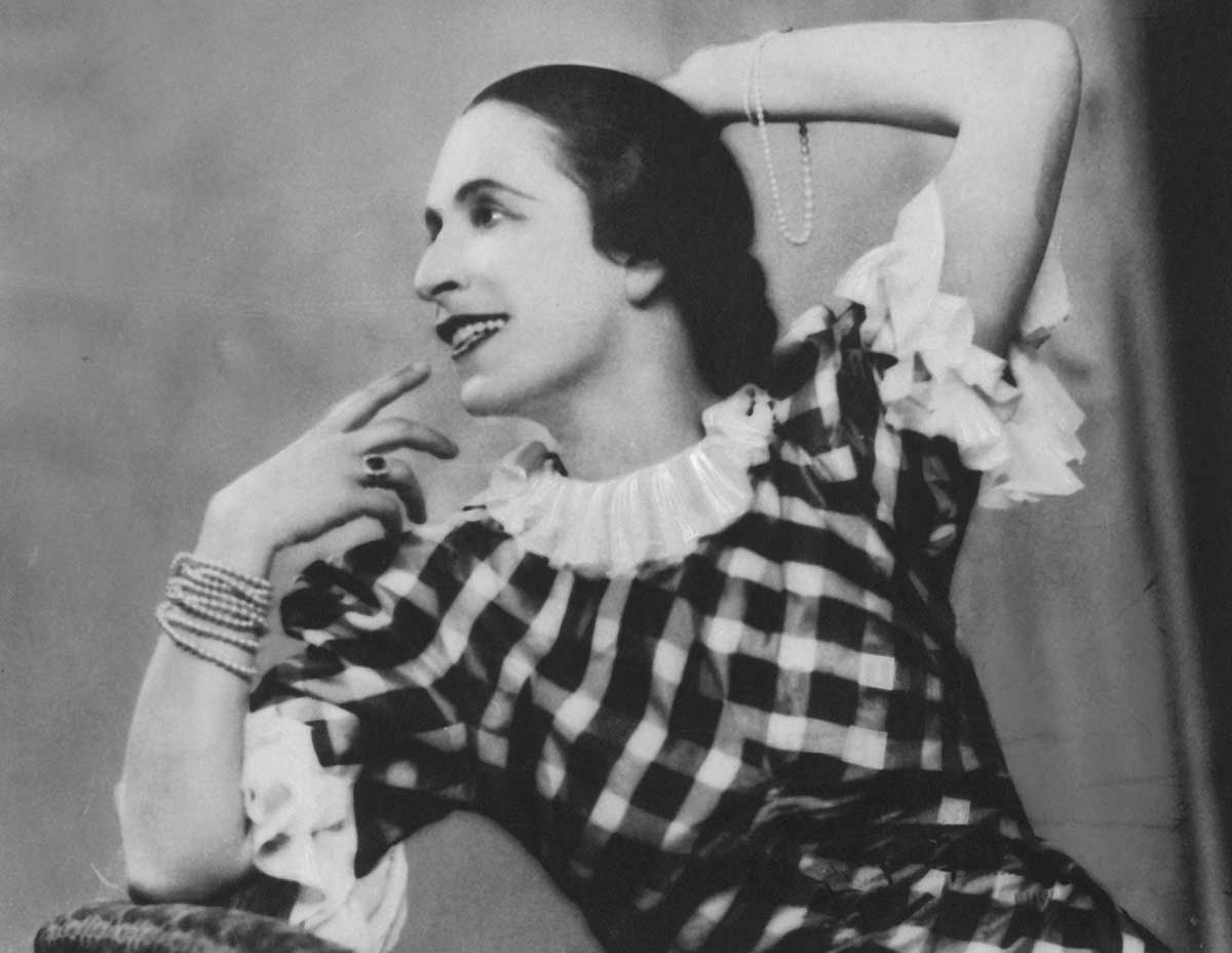 Black-and-white photograph of Gertrud Bodenwieser in a checkered dress, with ruffles around the neckline and on the sleeves, which sit just above her elbows. She is sitting, leaning on her right elbow and looking to the right. Her left arm is raised behind her head.