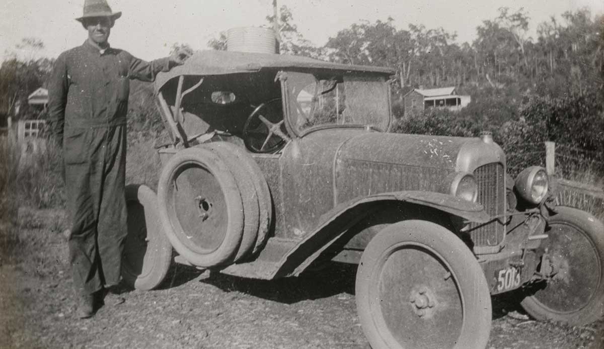 A black and white photograph of a man standing next to an automobile with his arm resting on the soft top roof. It is heavily mud-spattered and has a spare tyre fixed to the driver’s side and a number plate that reads ‘5013’. There are buildings in the distance.