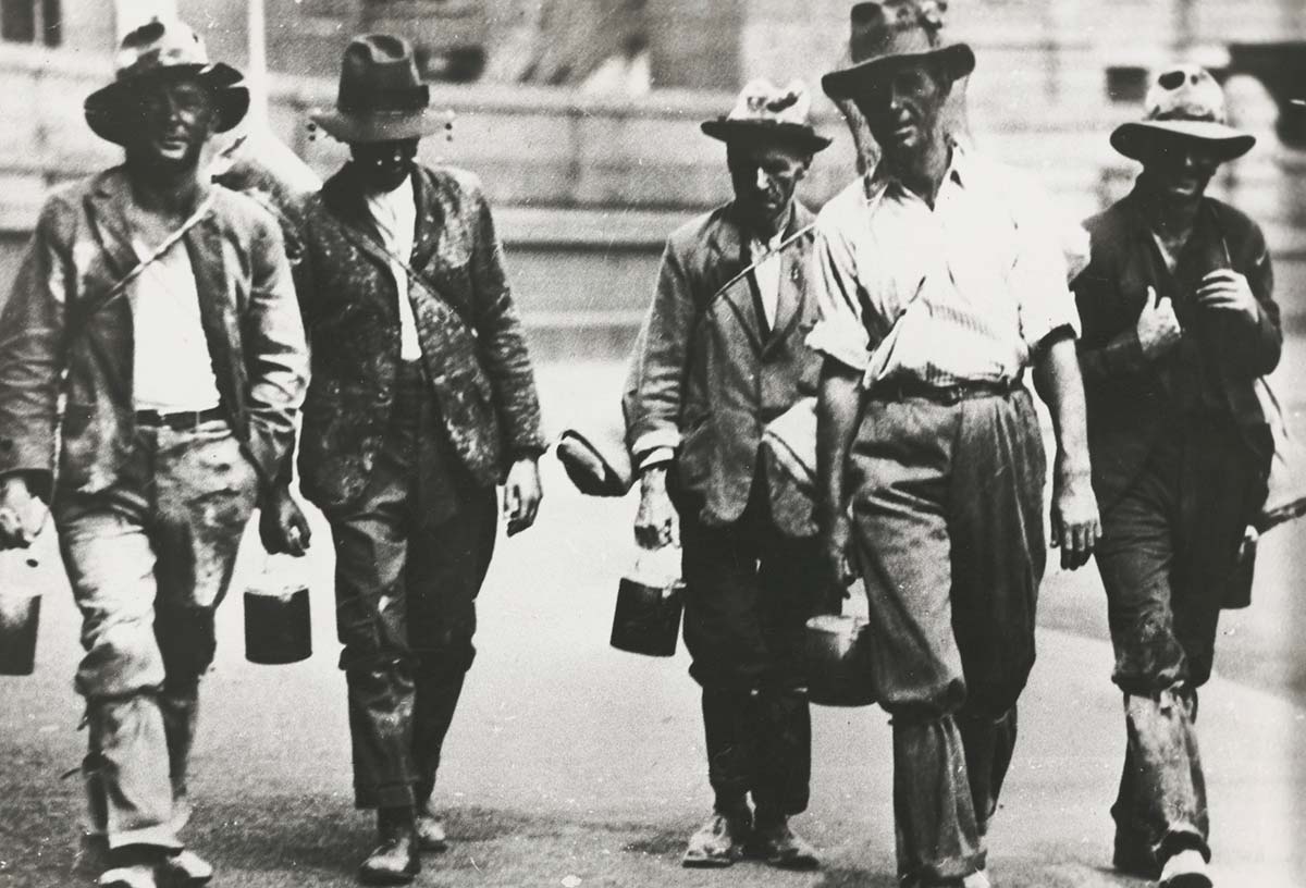 Five men walking towards the camera dressed in shabby clothes. Most are carrying swags and bill cans.
