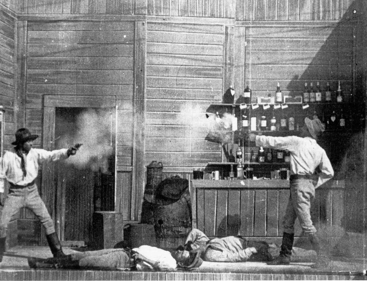 A shooting duel scene from a black and white movie, with two men firing at each other and two lying on the ground..