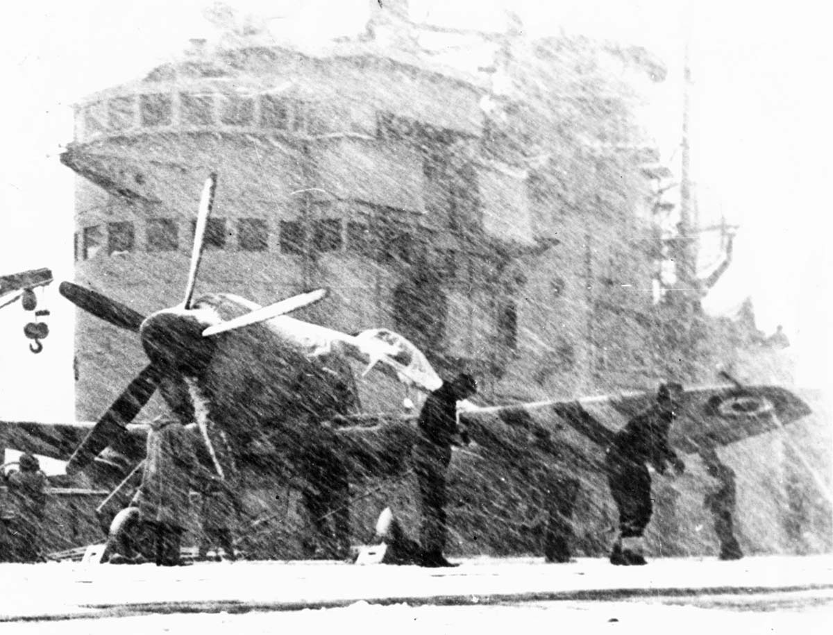 A fighter plane on the snow-covered deck of an aircraft carrier attended to by crew, with snow blowing hard.