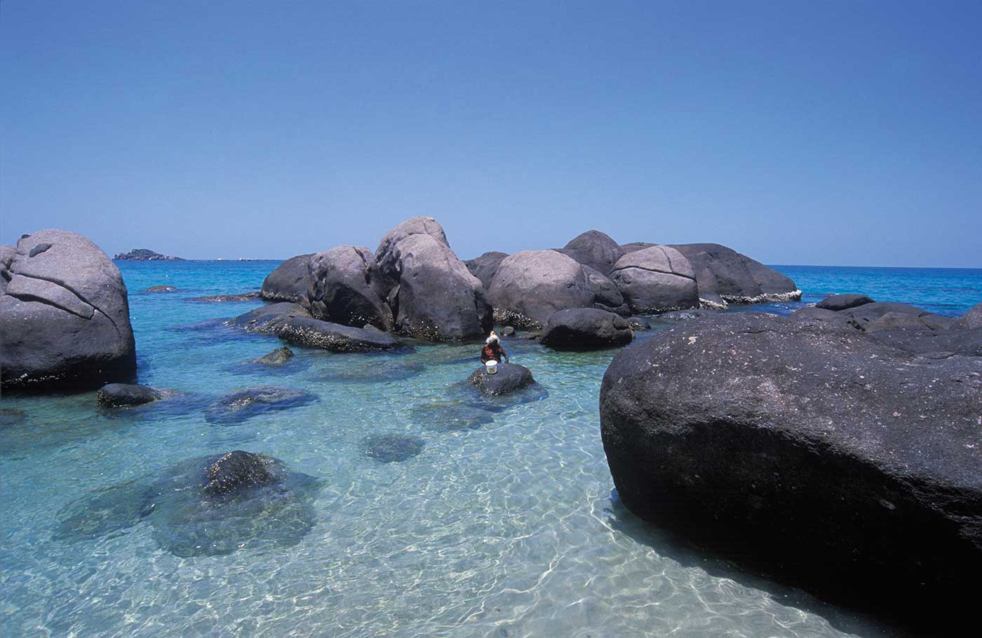 A colour photograph of a person wading in shallow water near a beach. Large rounded boulders surround the person. Submerged rocks can be seen in the clear blue water. Parts of the horizon are visible beyond the large boulders. The blue cloudless sky is in the top half of the photograph.