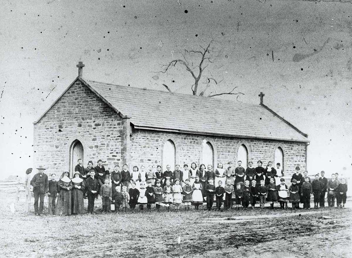 Faded black and white photo of a group portrait of children and staff standing in front of small stone church.