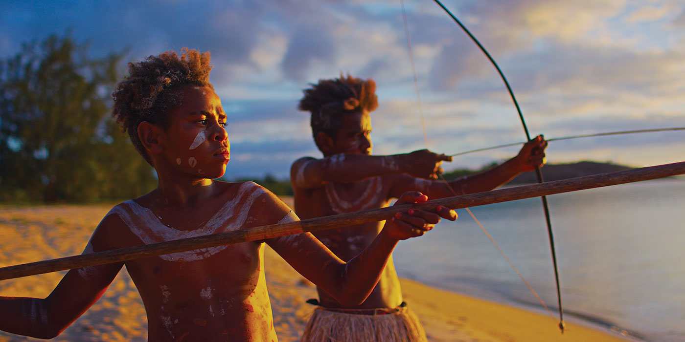 Two young boys are standing on a beach with spear and bow and arrow raised in attack mode.