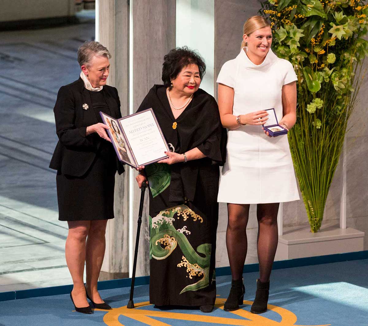 Three women pose for a photo at a ceremony. Two women are both holding up a double frame containing a certificate and a photo. The other woman is displaying a medal in an open box.