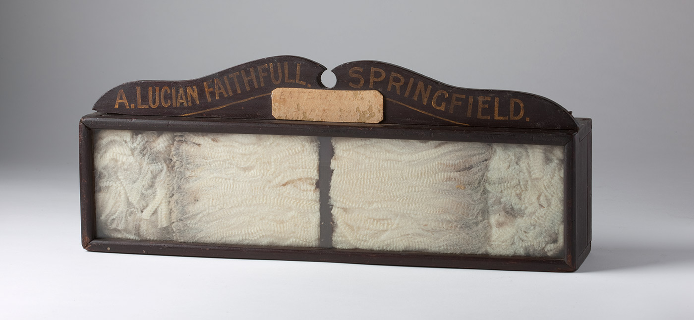 Glass-fronted wooden box, displaying a wool sample. Painted on a wooden banner at the top of the box: