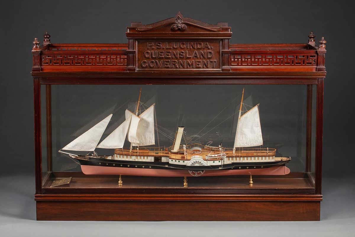 A model of a ship in a wood and glass case, the ship has five raised sails, water wheels on either side, painted in black and brown with details in blue and red, held on four gold coloured stands, and a gold plaque at one end. Th case is in good condition and has an engraved panel with the text:
