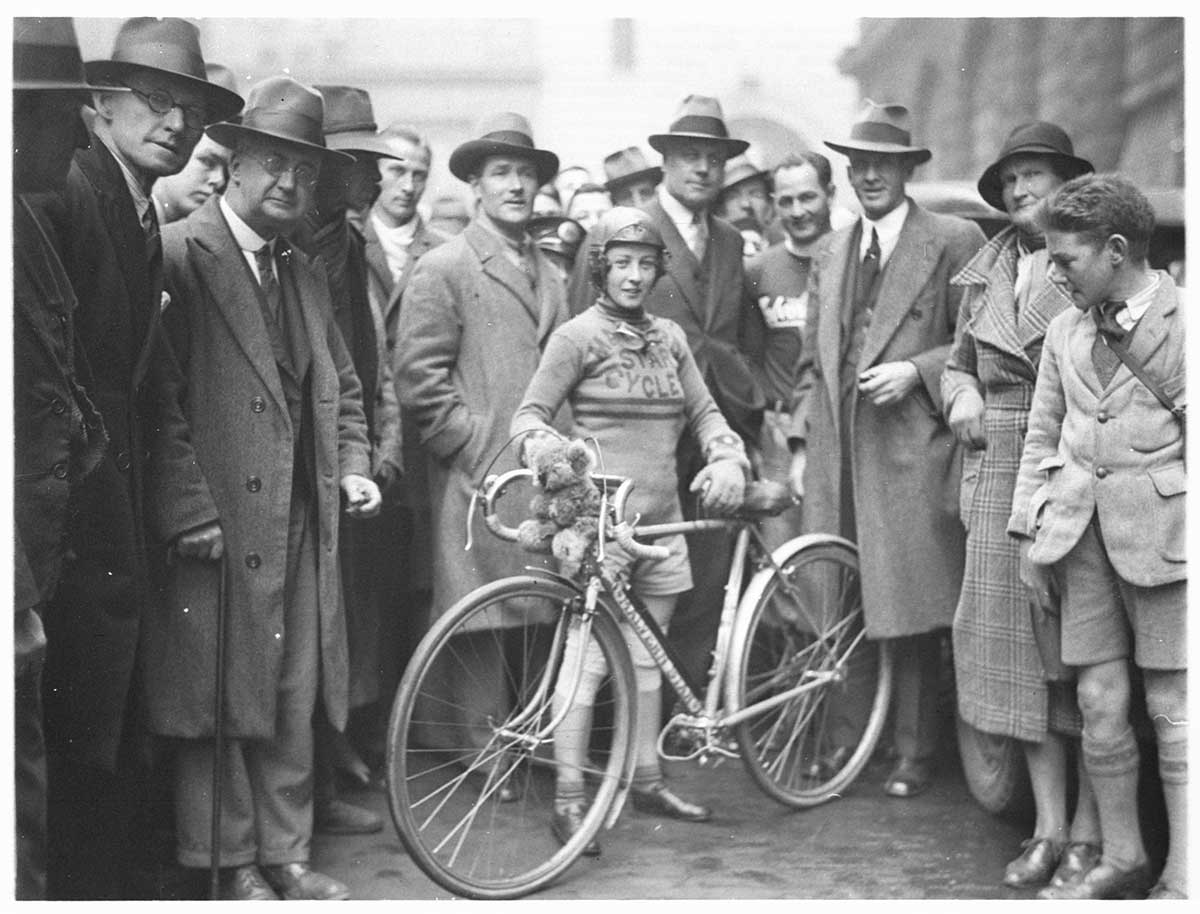 A black and white photo of Billie Samuels with her bike and surrounded by a crowd of people.