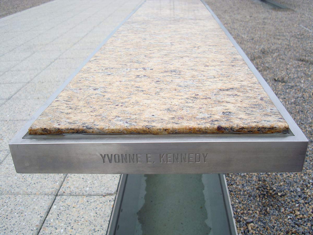 Far end of a silver rectangular metallic bench with a light marble top. 'YVONNE E. KENNEDY' is engraved in the side of the bench, which sits above a pool of water.